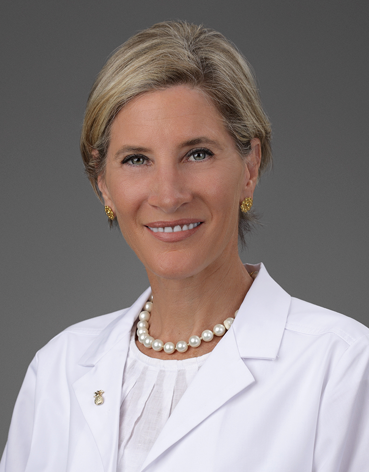 A professional headshot of a woman with short blonde hair, she smiles at the camera, she has on a white coat and pearls